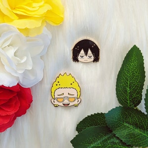 May include: Two gold-colored enamel pins featuring anime characters. The first pin depicts a character with black hair and glasses, while the second pin depicts a character with yellow hair and glasses. Both pins have a white background.