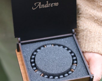 May include: A black wooden box with a black beaded bracelet inside. The box has a brown wood trim and the name "Andrew" engraved on the lid.