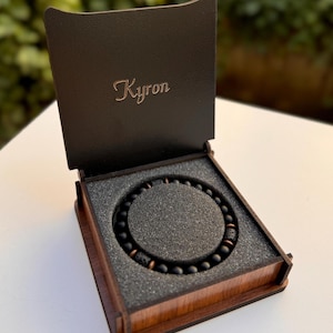 Peut inclure: Un bracelet de perles noires avec des accents cuivrés est présenté dans une boîte en bois avec le nom "Kyron" gravé sur le couvercle. La boîte est ouverte, révélant le bracelet niché dans un insert en mousse grise.