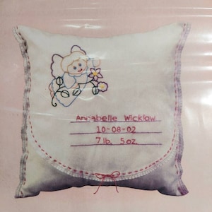 May include: A white pillow with pink stitching and a pink bow. The pillow has embroidered details of a baby angel with flowers and the text "Annabelle Wicklow 10-08-02 7 lb. 5 oz."