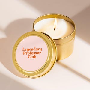 May include: A lit candle in a gold-colored tin with a lid. The lid has a pink label with the text "Legendary Professor Club" in orange. The candle is white, and the flame is yellow.