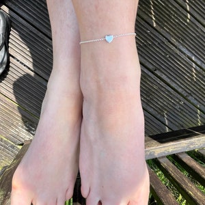 May include: A pair of bare feet with pink nail polish on the toes. The feet are resting on a wooden surface with green grass visible below. A silver heart-shaped anklet is worn on the right ankle.