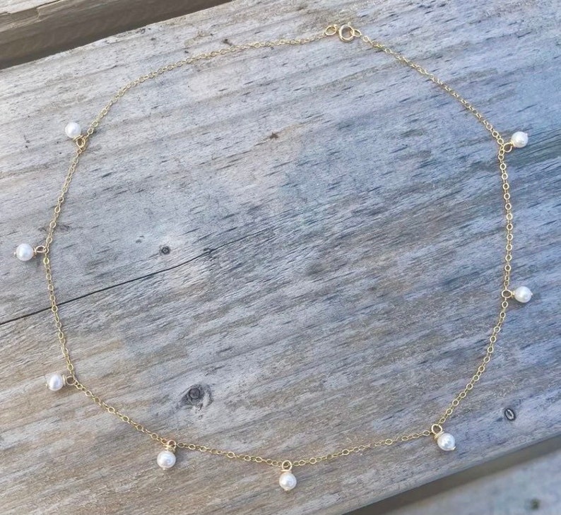 K&ouml;nnte beinhalten: Eine goldene Kettenkette mit kleinen wei&szlig;en Perlen, die daran h&auml;ngen. Die Kette liegt auf einer Holzoberfl&auml;che.