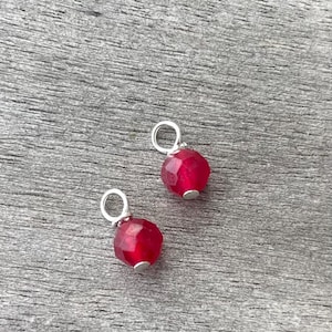 Può includere: Due piccoli ciondoli, ciascuno composto da una perla rotonda sfaccettata di colore rosso intenso fissata a un semplice anellino di metallo argentato. Le perle hanno una qualità traslucida e una superficie sfaccettata che riflette la luce.