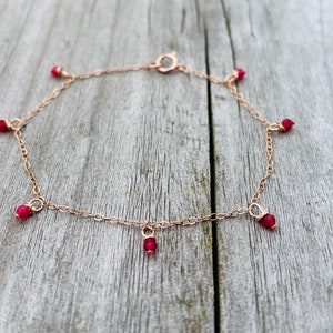 May include: A delicate gold chain bracelet with dangling red gemstone beads. The bracelet has a lobster clasp closure.