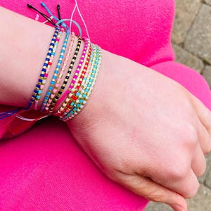 May include: A selection of colourful beaded bracelets, featuring shades of blue, pink, black, and gold. The bracelets are stacked on a wrist, displaying the diverse designs and colour combinations. The beads are small and uniform in size.