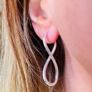 May include: A silver infinity hoop earring is worn in an ear. The earring is a continuous loop, shaped like the mathematical symbol for infinity. The earring is smooth and shiny, reflecting light. The background shows blonde hair and skin.