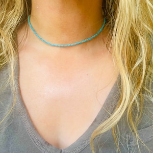 Puede incluir: Un delicado collar gargantilla de cuentas turquesa que lleva una persona. El collar está hecho de pequeñas cuentas redondas y es muy fino.