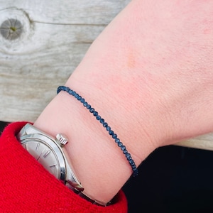 May include: A simple bracelet made of dark blue beads on a person's wrist. The bracelet is worn above a silver watch.