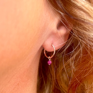 May include: A close-up of a person's ear wearing a small gold hoop earring with a dangling pink gemstone.