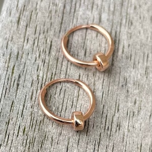 May include: Two rose gold hoop earrings with a small, rectangular accent. The earrings are displayed on a weathered, gray wooden surface. The hoops are small and simple, suitable for everyday wear.