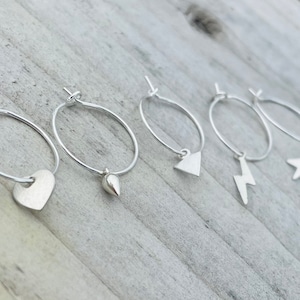 May include: Five silver hoop earrings with different charms. The charms include a heart, a teardrop, a triangle, a lightning bolt, and a star.