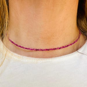May include: A delicate choker necklace composed of small, round, deep pink beads. The necklace is worn around the neck, resting on a white t-shirt. The beads appear to be evenly spaced, creating a simple yet elegant design.
