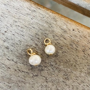 May include: Two small, gold-coloured charm pendants, each featuring a white pearl. The pendants are displayed on a wooden surface. The pearls have a round shape and are attached to the gold loops.