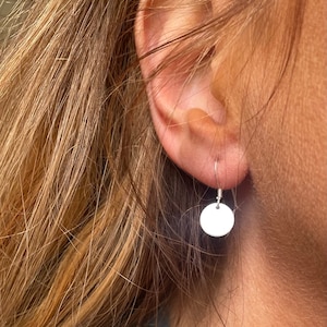 May include: A silver circle earring with a simple design. The earring is hanging from a person's ear.