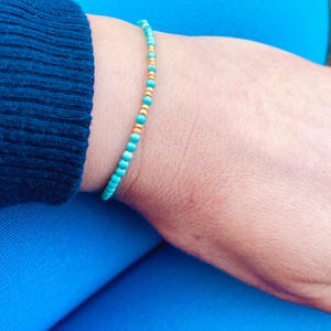 Op de afbeelding: Een turquoise en gouden kralen armband. De armband is gemaakt van kleine, ronde kralen die aan elkaar geregen zijn op een dun koord.