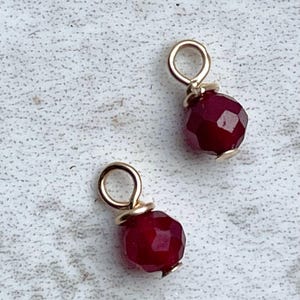 May include: Two gold-toned charms with a red gemstone bead. Each charm has a small loop for attaching to a necklace or bracelet.