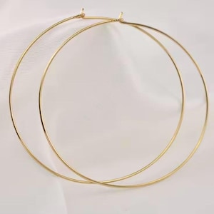 May include: A pair of large, gold-colored hoop earrings. The earrings are circular and appear to be made of a thin metal. They are displayed against a soft, white background, highlighting their simple design and metallic sheen.