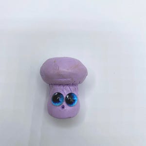 May include: A whimsical, handmade purple mushroom figurine with large, expressive blue eyes. The mushroom cap is a lighter shade of purple than the stem, creating a contrasting effect. The figurine is set against a plain white background.