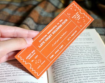 Op de afbeelding: Oranje boekenlegger met een ticketontwerp. Er staat "Late Night Readers Club" en "Levenslang lidmaatschap" op. De boekenlegger heeft een halve maan en sterren.