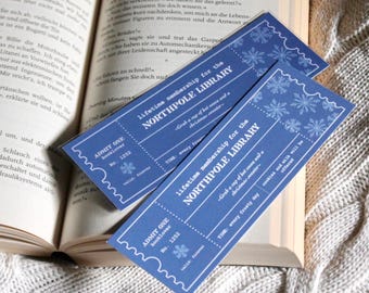 Op de afbeelding: Twee blauwe en witte ticketvormige bladwijzers met sneeuwvlokontwerpen. De bladwijzers luiden "Levenslange lidmaatschap voor de Noordpoolbibliotheek" en bevatten de tekst "Pak een kop warme chocolademelk en een kersttrui." en "Tijd: elke vriesdag".