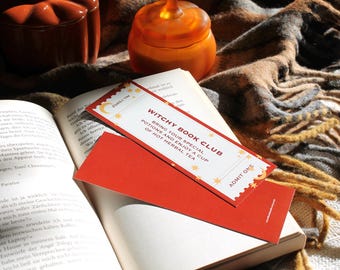 Op de afbeelding: Een open boek met een rode bladwijzer met de tekst "Witchy Book Club". De bladwijzer ligt bovenop een rode kaart. Twee oranje pompoenvormige containers staan op de achtergrond, samen met een bruine en grijze deken.