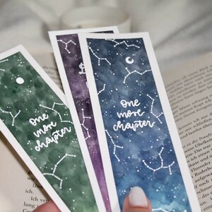 Könnte beinhalten: Drei Aquarell-Lesezeichen mit einem Galaxie-Design und weißen Sternbildern. Jedes Lesezeichen trägt den Text "One more chapter" in weiß.
