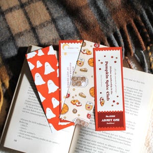 Op de afbeelding: Een stapel kleurrijke bladwijzers bovenop een open boek. De bladwijzers hebben verschillende ontwerpen, waaronder geesten, gebak en de tekst "Pumpkin Spice Club". De achtergrond omvat een geruite deken en een zacht, wit oppervlak.