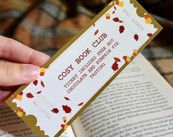 Op de afbeelding: Een gouden en witte bladwijzer met een ticketontwerp. De bladwijzer heeft de tekst "Cosy Book Club" en "Ticket includes free hot chocolate and pumpkin pie tasting". De bladwijzer is versierd met herfstbladeren en snoepmais.