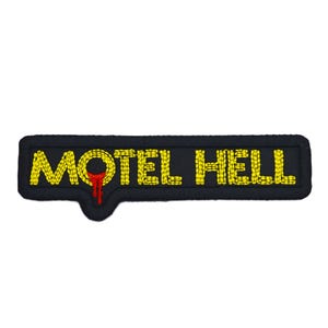May include: A black embroidered patch with the words "MOTEL HELL" in yellow thread. A red, blood-like drip design is below the "O" in "MOTEL". The patch has a rectangular shape with rounded edges.