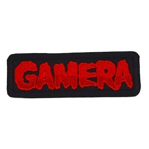 May include: A rectangular embroidered patch with a black background and a thick black border. The word GAMERA is stitched in bold, textured red thread across the centre, featuring a jagged, stylised font with a distinct, raised appearance.