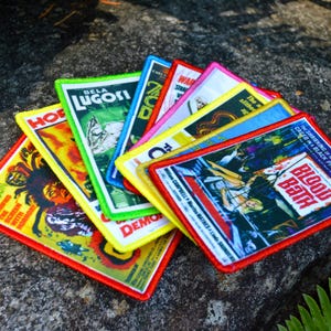 May include: A collection of colorful, rectangular patches featuring vintage horror movie posters. Each patch has a different design, with titles like "Blood Bath" and "Bela Lugosi." The patches are edged in various colors, including red, yellow, and green.