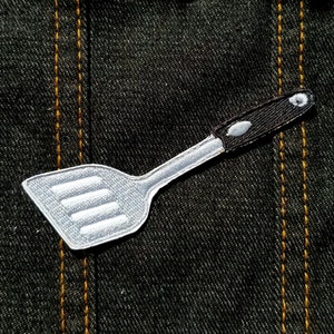 Peut inclure: Une spatule argentée avec un manche noir, brodée sur un tissu denim foncé.