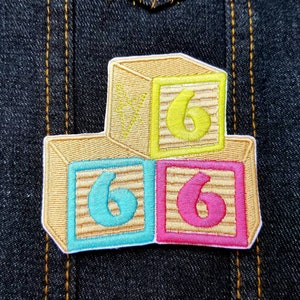 May include: Three wooden toy blocks stacked on top of each other. The top block is yellow with the number 6 on it. The middle block is blue with the number 6 on it. The bottom block is pink with the number 6 on it.