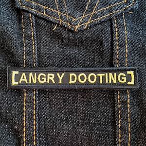 Könnte beinhalten: Eine schwarze Jeansjacke mit einem gelben gestickten Aufnäher, auf dem "[ANGRY DOOTING]" steht.