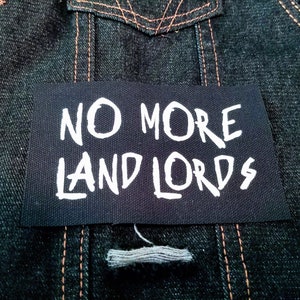May include: A black patch sewn onto a denim jacket with white text that reads "NO MORE LANDLORDS".