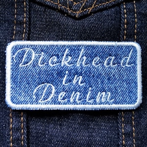 Może przedstawiać: Naszywka z niebieskiego denimu z białymi szwami i napisem "Dickhead in Denim" białym, ozdobnym pismem.