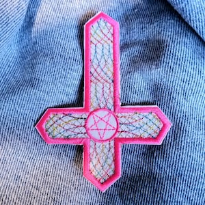 May include: Pink embroidered patch with a white pentagram inside a colorful web design. The patch is shaped like an upside-down cross.