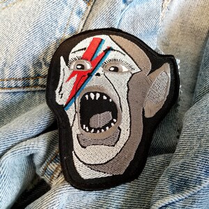 Puede incluir: Un parche bordado en blanco y negro de un monstruo gritando con un rayo rojo y azul en la cara.