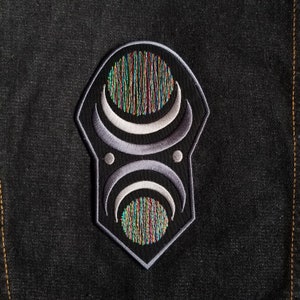 May include: A black and gray embroidered patch with a geometric design featuring a circle with concentric rings and two crescent moons.