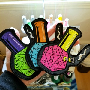 May include: Three embroidered patches featuring a bong and a 20-sided die. The patches are in green, blue, and pink with black outlines. The bongs are purple, orange, and yellow.