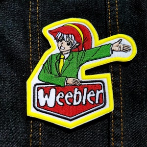 May include: A yellow, red, and green embroidered patch with a cartoon character wearing a red hat and green suit. The character is holding a microphone and the text "Weeble" is written on the patch.