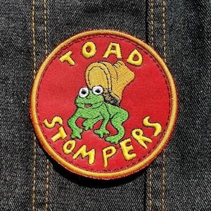 May include: A red and yellow embroidered patch with a cartoon frog wearing a hat. The text "TOAD STOMPERS" is embroidered around the frog.