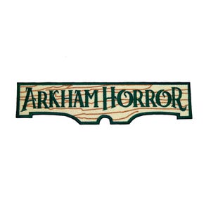 May include: Embroidered patch featuring the text "ARKHAM HORROR" in dark green lettering. The patch has a light beige background with brown wood grain lines, and a dark green border. The patch has a unique shape with a cutout at the bottom.