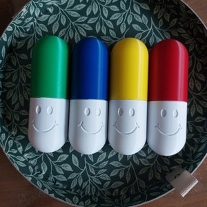 Puede incluir: Cuatro contenedores con forma de píldora con caras sonrientes en la parte delantera. Los contenedores son verde, azul, amarillo y rojo.