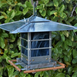 May include: A handcrafted bird feeder with a pagoda-style roof and a square body. The feeder is made of metal with a blue-gray finish and features clear glass panels with a smoky blue design. The base has a wooden perch.