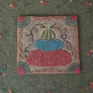 May include: A hand-stitched rug with a colorful pumpkin design. The rug is made with a variety of textures and colors, including brown, red, blue, and yellow. The rug is surrounded by a green background with a vine pattern.