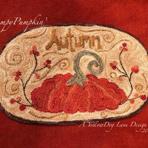 May include: A beige oval rug with a brown border, featuring a red and orange pumpkin with a green stem and the word "Autumn" in brown lettering. The rug is decorated with brown vines and red berries. The text "Lumpy Pumpkin" and "A Yellow Dog Lane Design 2011" are also included.