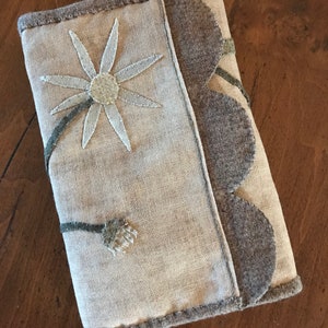 Può includere: Una copertina per libro in tessuto fatta a mano con un motivo di margherita bianca e un bordo in feltro marrone. La copertina è in tessuto di lino beige chiaro.