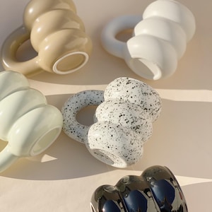 Op de afbeelding: Een assortiment keramische mokken in verschillende kleuren. De mokken hebben een uniek, afgerond ontwerp met een grote handgreep. Kleuren zijn beige, wit, mintgroen, gespikkeld zwart en wit en glanzend zwart. De mokken zijn geplaatst op een lichtgekleurd oppervlak.